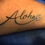 Фото татуировки надписи 'Aloha' на предплечье. Черно-белое изображение, выполненное в каллиграфическом стиле.