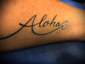 Фото татуировки надписи 'Aloha' на предплечье. Черно-белое изображение, выполненное в каллиграфическом стиле.