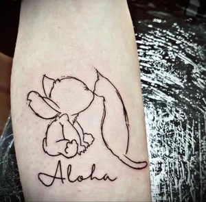 Фото татуировки надписи 'Aloha' под изображением Стича из мультфильма Lilo & Stitch. Черно-белый рисунок на руке.