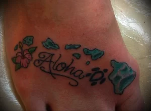 Фото татуировки надписи 'Aloha' на стопе, украшенной цветами гибискуса и бабочками. Цветная татуировка в ярких тонах.