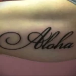Фото татуировки надписи 'Aloha' с декоративными завитушками. Черно-белое изображение на коже, выполненное в каллиграфическом стиле.