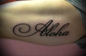 Фото татуировки надписи 'Aloha' с декоративными завитушками. Черно-белое изображение на коже, выполненное в каллиграфическом стиле.