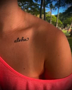 Фотография татуировки надписи 'Aloha' на плече девушки. Небольшая, изящная татуировка черного цвета на загорелой коже.