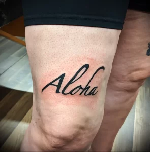 Черно-белое фото татуировки надписи 'Aloha' на колене. Минималистичный дизайн, четкие линии, небольшое изображение.