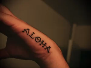 Фото тату на пальце с надписью 'Aloha', выполненное в черном цвете тонкими линиями. Идеально для любителей небольших и стильных татуировок.