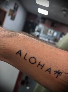 Фото тату на руке с надписью 'Aloha' и небольшим листиком, выполненное в черном цвете тонкими линиями. Минималистичный и стильный дизайн.