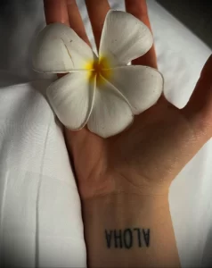 Фото тату на запястье с надписью 'Aloha', выполненное в черном цвете тонкими линиями. Идеально для любителей простых и элегантных татуировок.