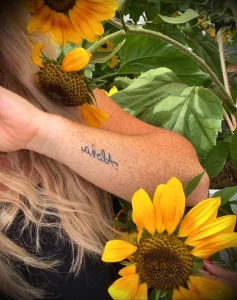 Фото тату на руке с надписью 'Aloha', расположенной на фоне ярких подсолнухов, выполненное в мягких тонах.