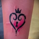 Фото тату на руке в стиле Kingdom Hearts с изображением черного сердца с ключом и розовыми брызгами краски. Стиль татуировки - минимализм.