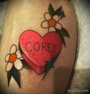Фото тату на бедре: красное сердце с надписью "Corey" внутри, окруженное белыми цветами и зелеными листьями.