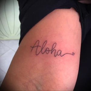 Фото тату с надписью 'Aloha' и маленьким сердечком на руке. Простой и элегантный дизайн, идеально подходит для тех, кто ценит минимализм.