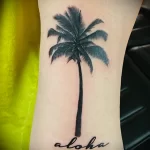 Фото тату пальмы с надписью 'Aloha' на руке. Минималистичный дизайн и черно-серые тона создают стильный и лаконичный образ.