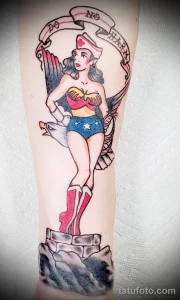 Фото тату Wonder Woman в классическом костюме с надписью 'Do No Harm'. Татуировка выполнена в стиле олд скул на руке.