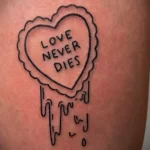 Фото тату на руке: черное сердце с надписью 'LOVE NEVER DIES'. Простой и лаконичный рисунок татуировки.