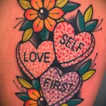 Фото тату на плече: три сердца с надписями 'Love Self First' и цветами в стиле олд скул. Яркие цвета и детальная проработка.