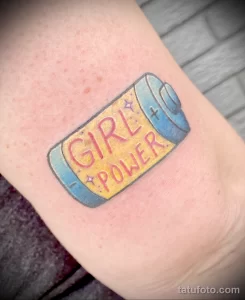 Фото яркой татуировки на руке с изображением батарейки и надписью 'Girl Power'. Использованы цвета: синий, розовый и желтый.