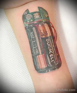 Фото реалистичной татуировки на предплечье с изображением батарейки Duracell и микросхемы. Использованы яркие цвета и детализированная прорисовка.