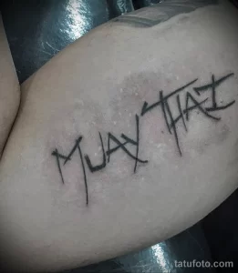 Фото тату на руке: надпись "MUAY THAI" в стиле brush lettering, выполненная в черном цвете.