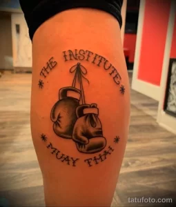 Фото тату на ноге: боксерские перчатки в круге с надписями 'THE INSTITUTE' и 'MUAY THAI'. Черно-белый рисунок татуировки.