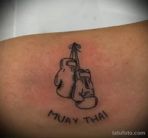 Фото тату на руке: контурное изображение боксерских перчаток и надпись 'Muay Thai'. Минималистичный рисунок татуировки.