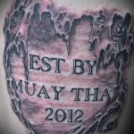 Фото татуировки Муай Тай на плече, выполненной в виде печати с надписью "EST BY MUAY THAI 2012" и изображением перчаток.
