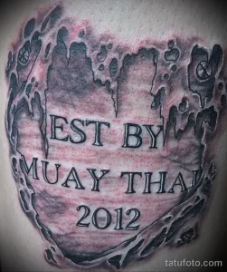 Фото татуировки Муай Тай на плече, выполненной в виде печати с надписью "EST BY MUAY THAI 2012" и изображением перчаток.