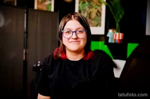Фото тату мастера, работающего над татуировкой. Девушка-тату мастер в очках и черной футболке, сосредоточенно смотрит на клиента.