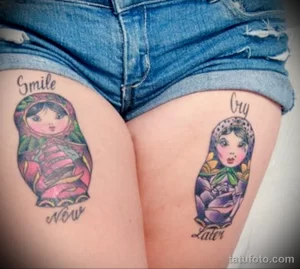 Фото тату на бедрах: две матрёшки с надписями "Smile Now" и "Cry Later". Яркие цвета и традиционный русский стиль.