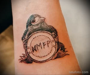 Фото тату птички, держащей в лапах круг с буквами 'MMFP'. Черно-белый рисунок татуировки выполнен в минималистичном стиле на предплечье.