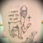 Фото тату с изображением странного существа с рогами и надписью "I don't want to be God" на ноге. Черно-белый рисунок, детализированная проработка.