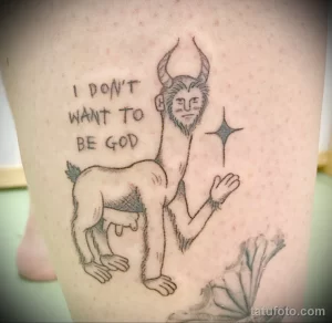 Фото тату с изображением странного существа с рогами и надписью "I don't want to be God" на ноге. Черно-белый рисунок, детализированная проработка.