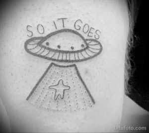 Фото тату с изображением НЛО и надписи "So it goes" на руке. Черно-белый рисунок, минималистичный стиль.