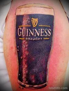 Фото татуировки на руке: стакан пива Guinness с логотипом и пузырьками. Реалистичный стиль тату, темные тона, тематика пива.