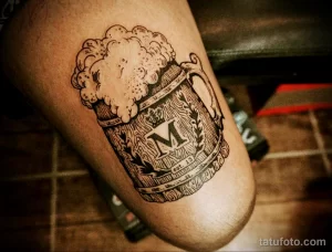 Фото тату кружки пива с логотипом 'M' и надписью 'est. 19...'. Черно-белый рисунок татуировки с высокой детализацией и текстурой.