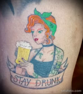 Фото тату с изображением рыжеволосой девушки с кружкой пива и надписью "Stay Drunk". Яркий рисунок в традиционном стиле.