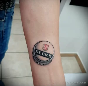 Фото татуировки на предплечье с изображением крышки от пива 'Beck's'. Черно-белый рисунок с реалистичной детализацией и логотипом бренда.