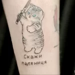 Небольшая татуировка на предплечье с изображением серой кошки, держащей нож, и надписью 'Скажи Паляница'. Необычный рисунок татуировки.