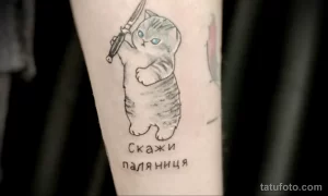 Небольшая татуировка на предплечье с изображением серой кошки, держащей нож, и надписью 'Скажи Паляница'. Необычный рисунок татуировки.