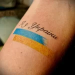 Фото тату с надписью "Из Украины" и изображением флага Украины на руке. Яркий рисунок с использованием синего и желтого цветов. Эскиз татуировки, символизирующий патриотизм.