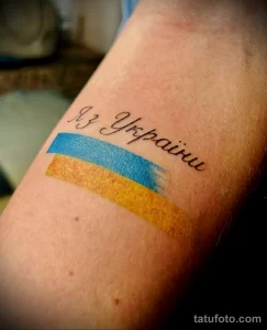 Фото тату с надписью "Из Украины" и изображением флага Украины на руке. Яркий рисунок с использованием синего и желтого цветов. Эскиз татуировки, символизирующий патриотизм.