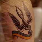 Фото тату на плече с изображением ласточки с лентой в клюве. Традиционный рисунок татуировки в стиле олдскул.