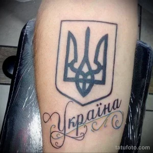 Фото татуировки на руке с изображением украинского трезубца и надписи 'Україна'. Черно-белый рисунок татуировки.