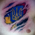 Фото тату украинского тризуба, словно прорывающегося сквозь разорванную кожу на груди. Рисунок татуировки в реалистичном стиле с использованием синих, желтых и красных цветов.