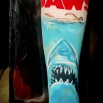 Фото тату на руке: гиперреалистичное изображение акулы из фильма "Челюсти", вырывающейся из воды. Детализированная татуировка с яркими цветами.