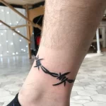 Фото тату: черная колючая проволока, обвивающая лодыжку. Простой и лаконичный рисунок татуировки, выполненный в графическом стиле.