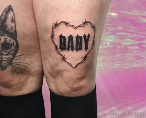 Фото татуировки колючей проволоки в форме сердца с надписью 'BABY' на колене. Черно-белый рисунок, выполненный в графическом стиле.