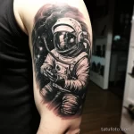 Фото тату: реалистичный рисунок космонавта на фоне звездного неба и галактик, выполненный в черно-серых тонах с высокой детализацией.