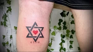 Фото тату на ноге мужчины, демонстрирующего минималистичную татуировку в форме звезды Давида с красным сердцем в центре и датой.