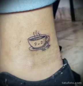 Фото тату маленькой чашки кофе с нарисованными глазками и улыбкой на лодыжке. Минималистичный рисунок татуировки в черных линиях.