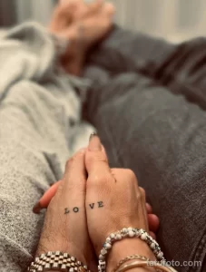 Фотография пары, держащейся за руки, на руках которых нанесены слова "LOVE" (перевернутое и обычное) черной краской. Татуировка выполнена в минималистичном стиле.